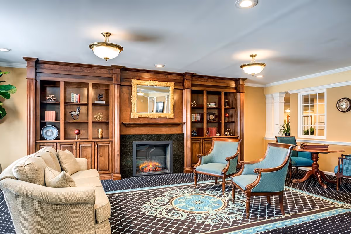 A cozy living room area featuring a beige sofa with cushions, two wooden armchairs with teal upholstery, and a patterned carpet with blue and beige designs. The room has a wooden built-in bookshelf and cabinet unit with decorative items and books, centered around a fireplace with a glowing fire and a large ornate gold-framed mirror above it. The walls are painted light yellow, and there are ceiling lights providing warm illumination. In the background, there is a small round wooden table with chairs and a window with white trim.
