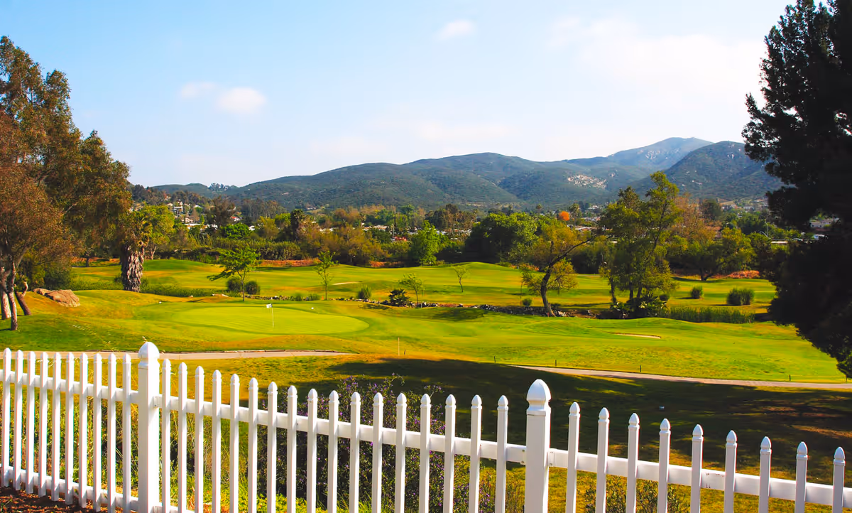 View of a green golf course with a white picket fence in the foreground, surrounded by trees and hills under a clear blue sky.