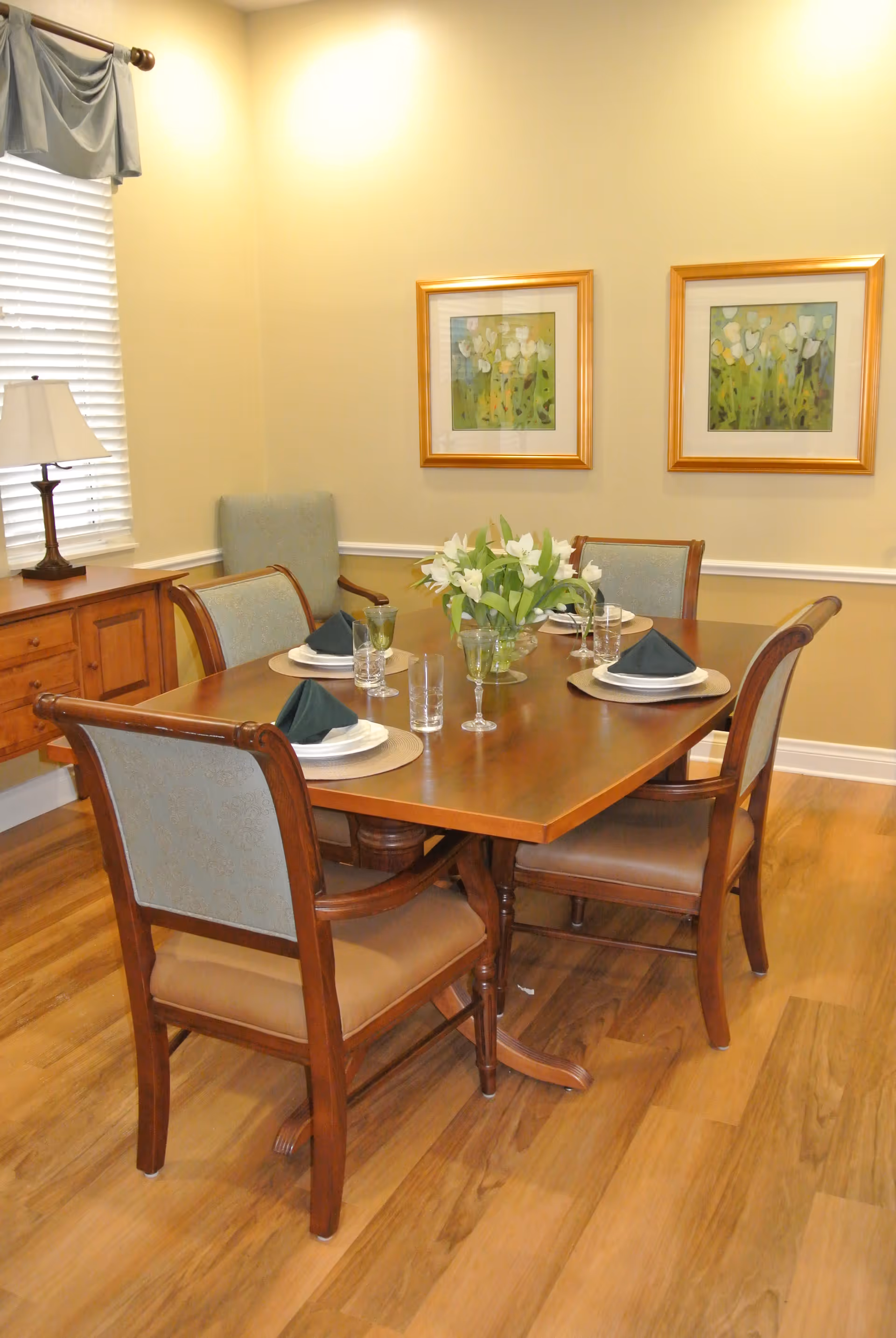 Dining room with a wooden table set for four, a floral centerpiece, upholstered chairs and framed wall art.