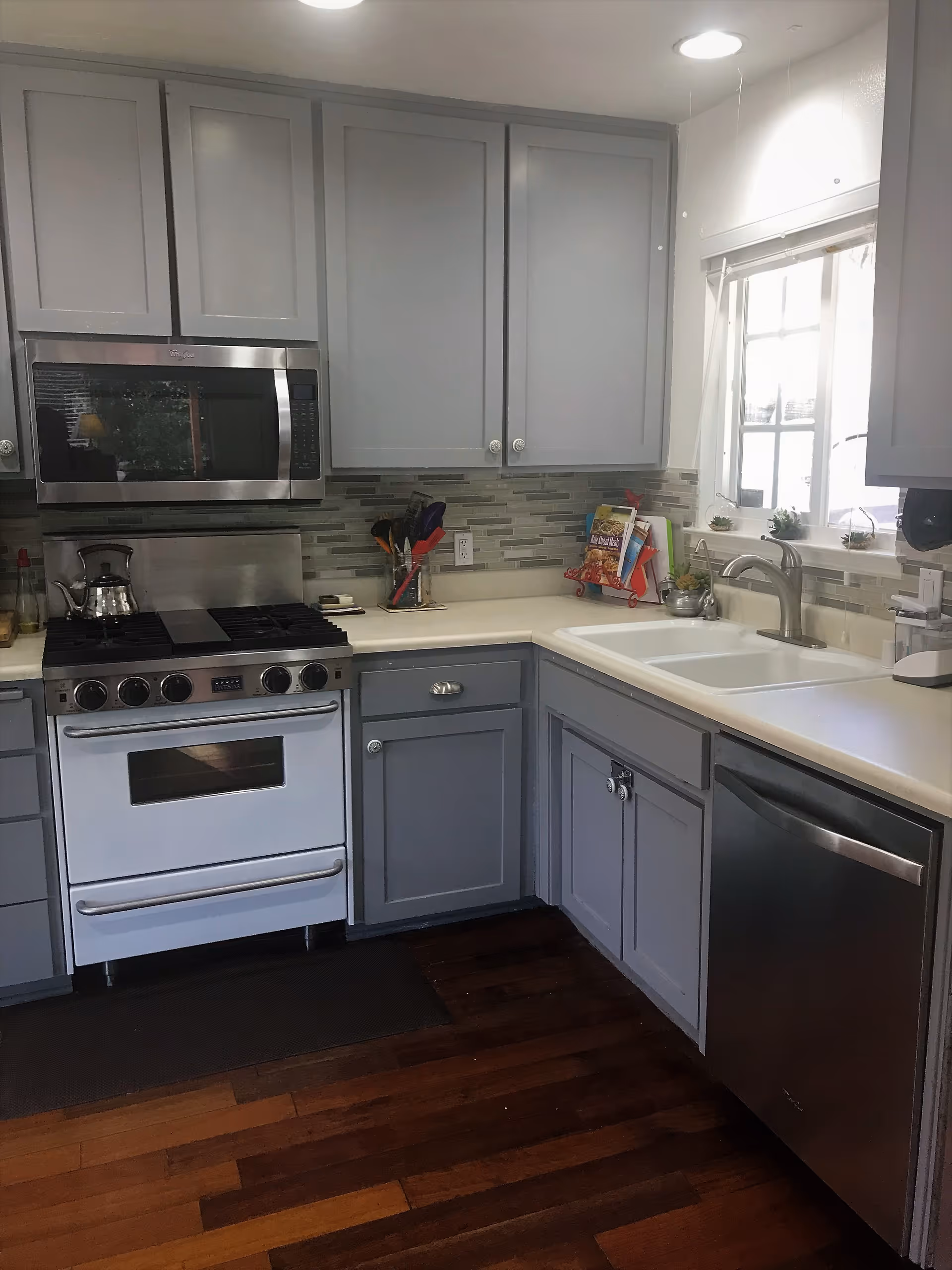 Bright modern kitchen with gray cabinets, stainless steel appliances including a gas range and microwave, and a double sink under a window.