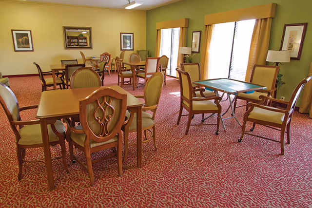 A well-lit room with multiple wooden tables and chairs arranged on a red patterned carpet. The walls are painted green and yellow, with framed artwork and windows with yellow curtains allowing natural light to enter. The room appears to be a communal or activity area with a calm and inviting atmosphere.
