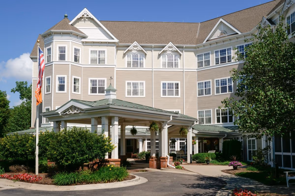 Front exterior of a multi-story assisted living building with a covered porte-cochère, flagpole, and landscaped entrance.