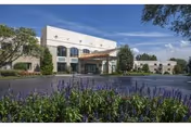 Exterior view of a senior living facility building with a covered entrance, surrounded by trees and landscaping including purple flowering plants in the foreground under a clear blue sky.