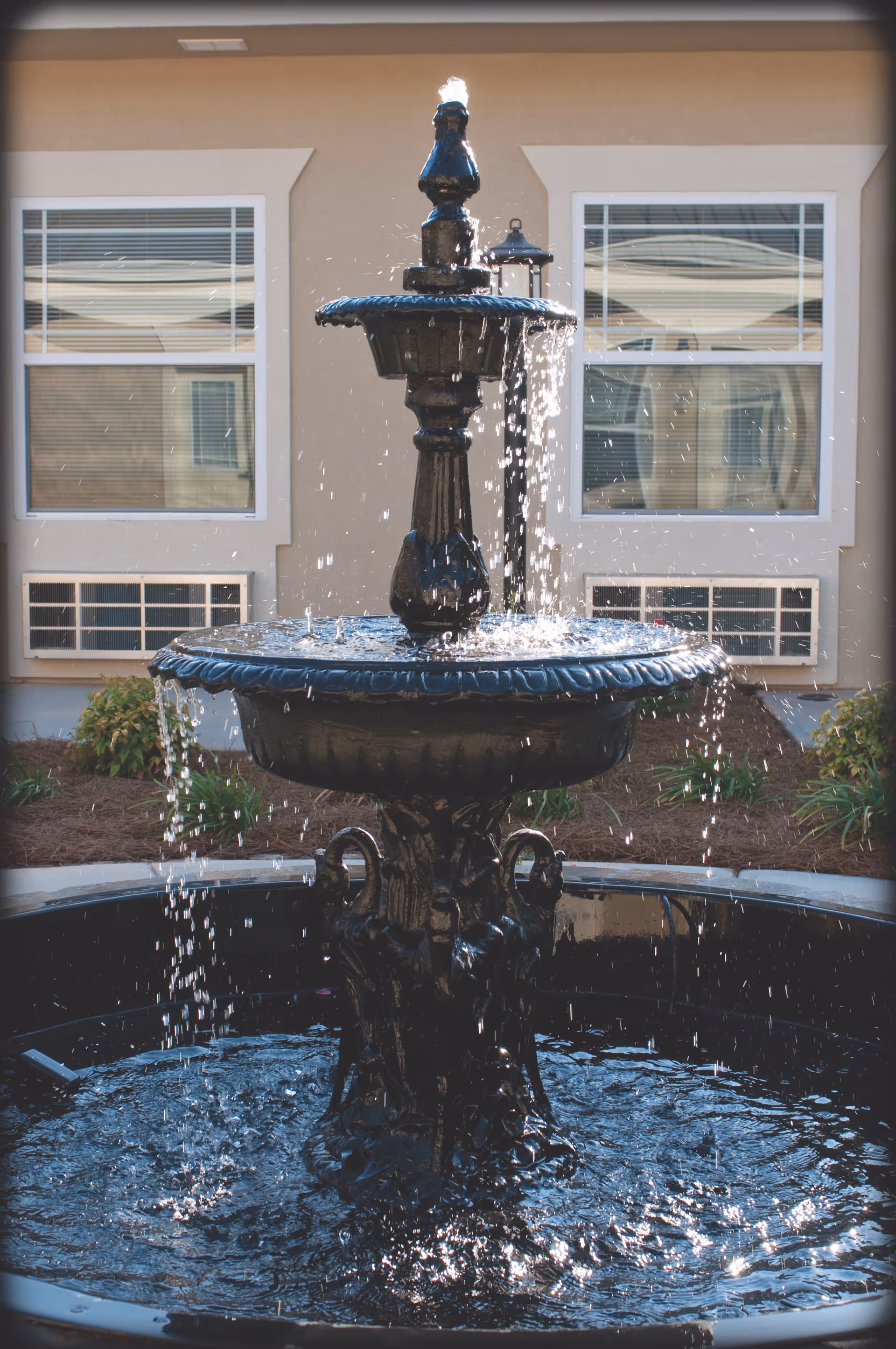 A decorative black water fountain with water cascading down multiple tiers, situated outdoors in front of a beige building with two windows and some landscaping including small bushes and mulch.