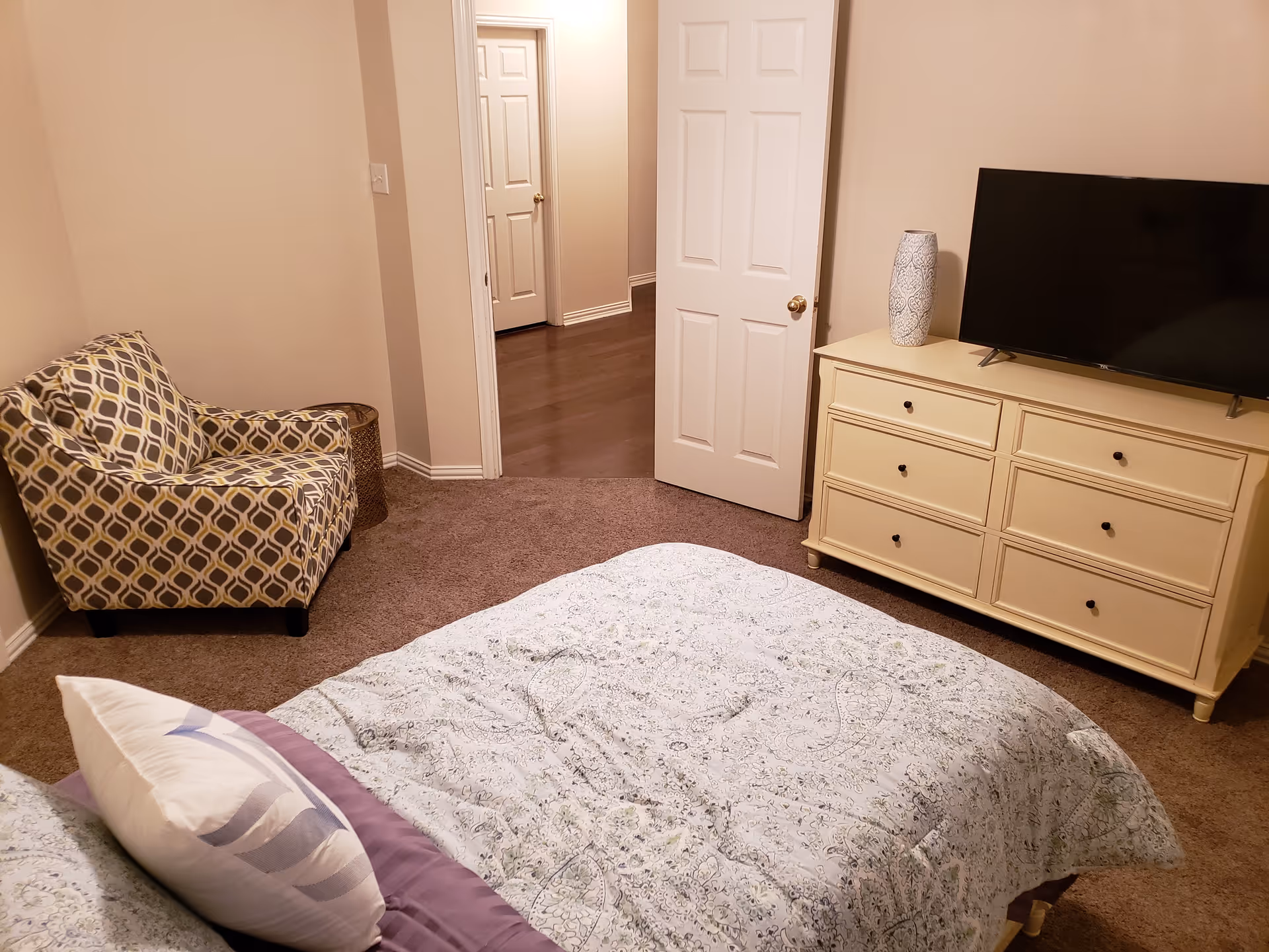 A bedroom with a bed covered in a patterned comforter and pillows, a patterned armchair in the corner, a cream-colored dresser with six drawers, a decorative vase on top, and a flat-screen TV. The door is open, showing a hallway with wooden flooring and another closed door.