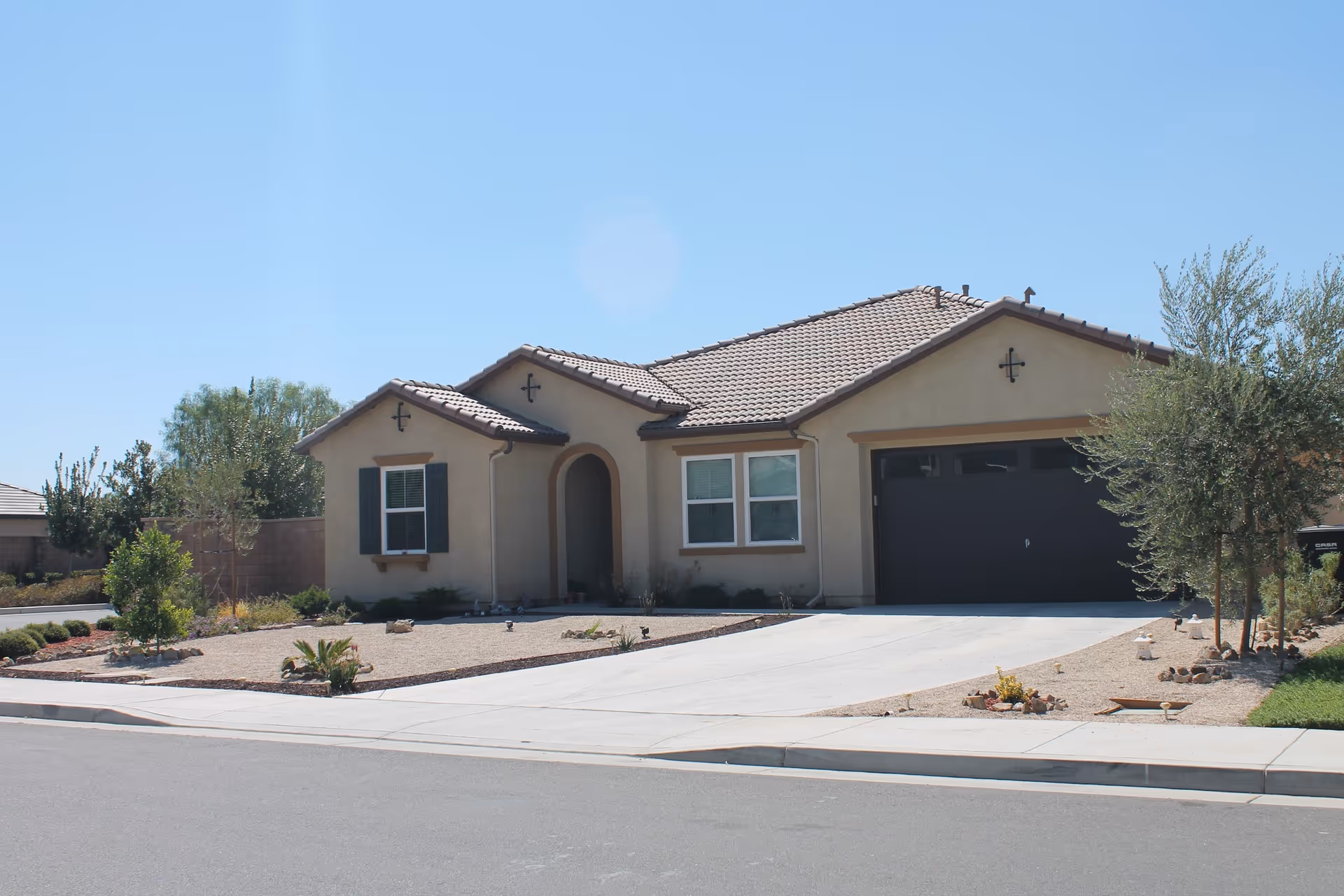 Single-story residential building with a tiled roof, beige exterior walls, a two-car garage with a dark door, and a driveway. The front yard has minimal landscaping with small plants and gravel. The sky is clear and blue.