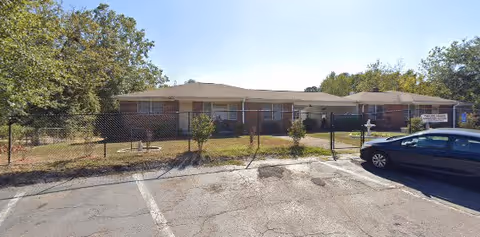 Single-story residential building with a fenced front yard, small trees, and a parked car in the parking lot under a clear sky.