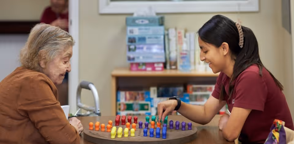 An elderly woman and a young woman sitting at a table playing a board game with colorful pieces in a cozy indoor setting with shelves and books in the background.