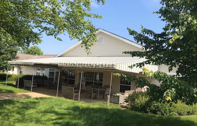 Exterior view of a single-story building with a covered patio area furnished with chairs and tables. The building is surrounded by green grass, trees, and shrubs under a clear blue sky.
