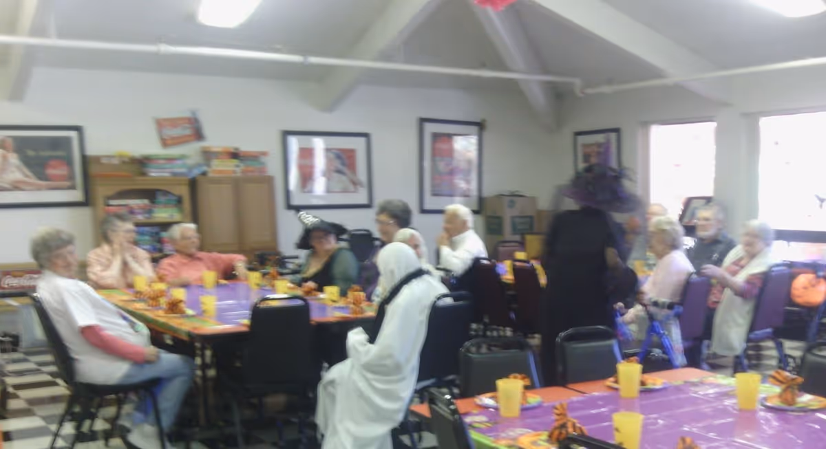 A group of elderly people gathered in a decorated common room, sitting around tables covered with purple tablecloths and orange-themed decorations, some wearing costumes including a witch hat and a ghost outfit.