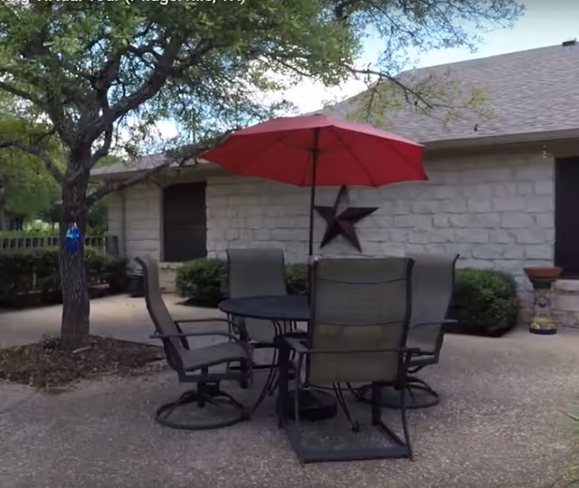 Outdoor patio area with a round table and four chairs under a red umbrella. The patio is adjacent to a stone building with a large star decoration on the wall. There is a tree nearby and some bushes along the building.
