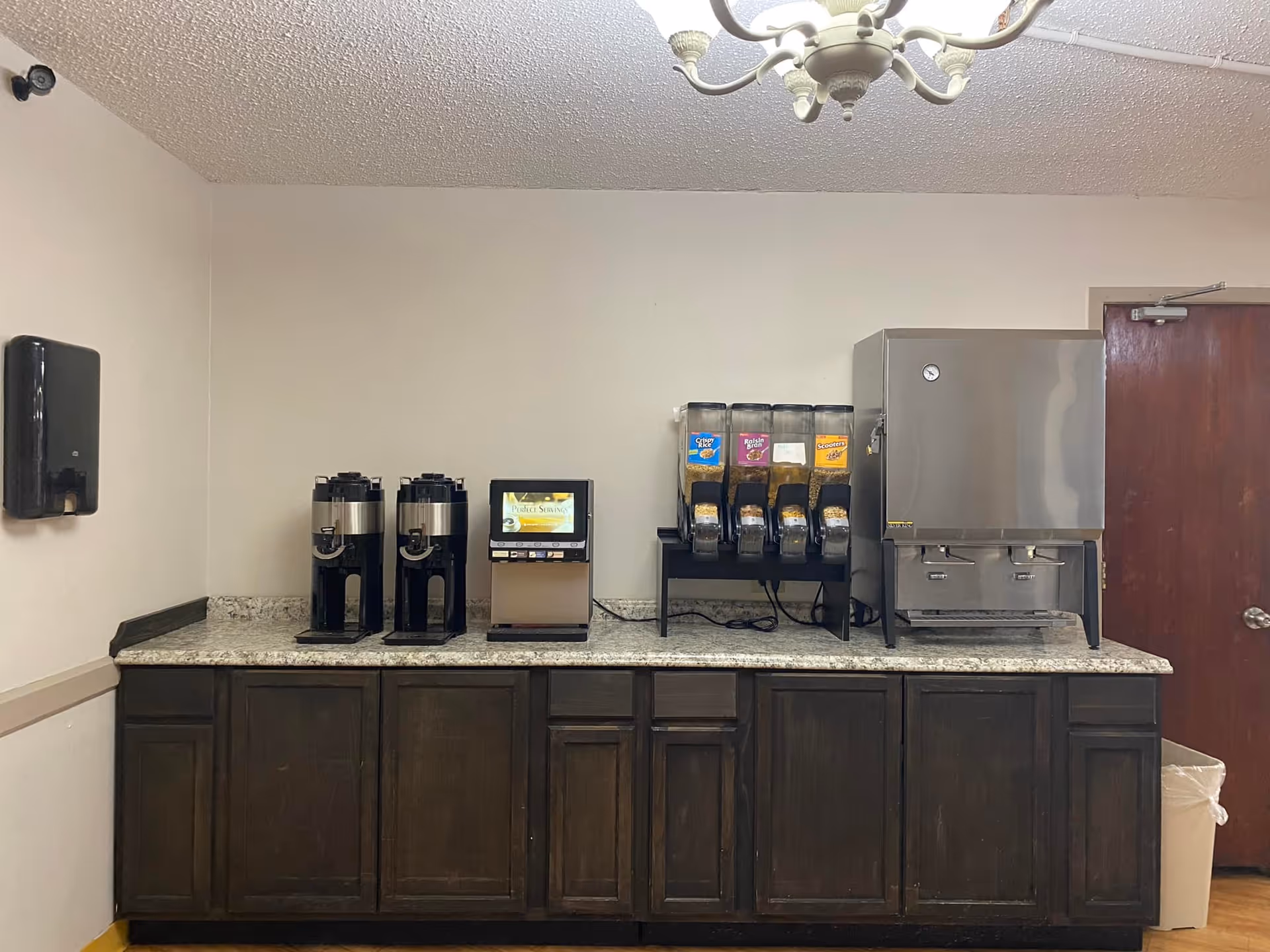A beverage station with two coffee dispensers, a hot water or tea dispenser, three cereal dispensers, and a large stainless steel water dispenser on a granite countertop with dark wooden cabinets below. There is a paper towel dispenser mounted on the left wall and a closed wooden door on the right side.