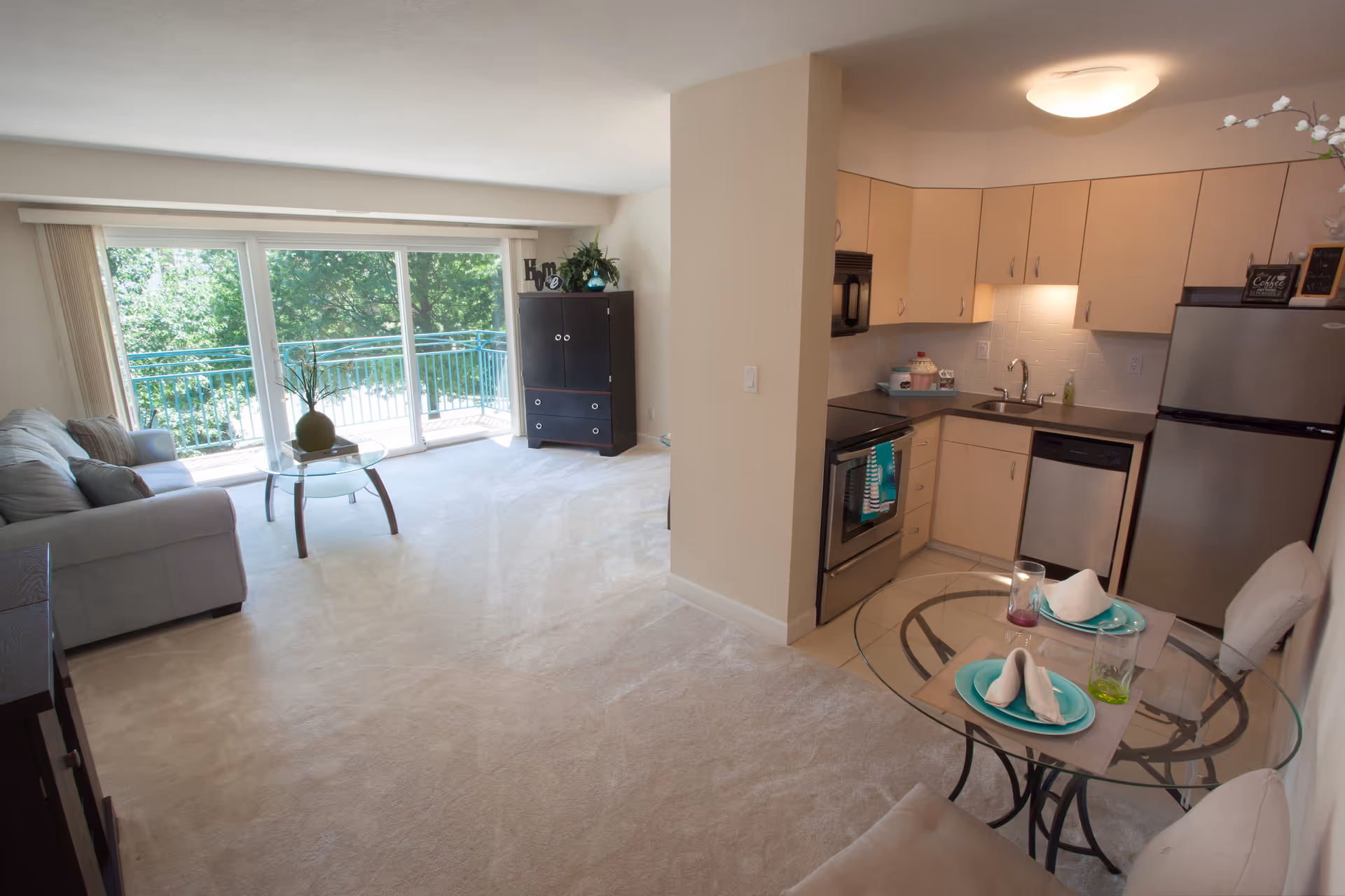 A bright and clean apartment interior featuring a small kitchen with light wood cabinets, stainless steel appliances, and a glass dining table set for two. The living area has a gray sofa, a glass coffee table with a decorative vase, and a dark wood cabinet. Large sliding glass doors open to a balcony with a green leafy view outside.