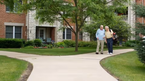 Two elderly men walking and talking on a paved pathway surrounded by green grass and trees in front of a brick and stone residential building.