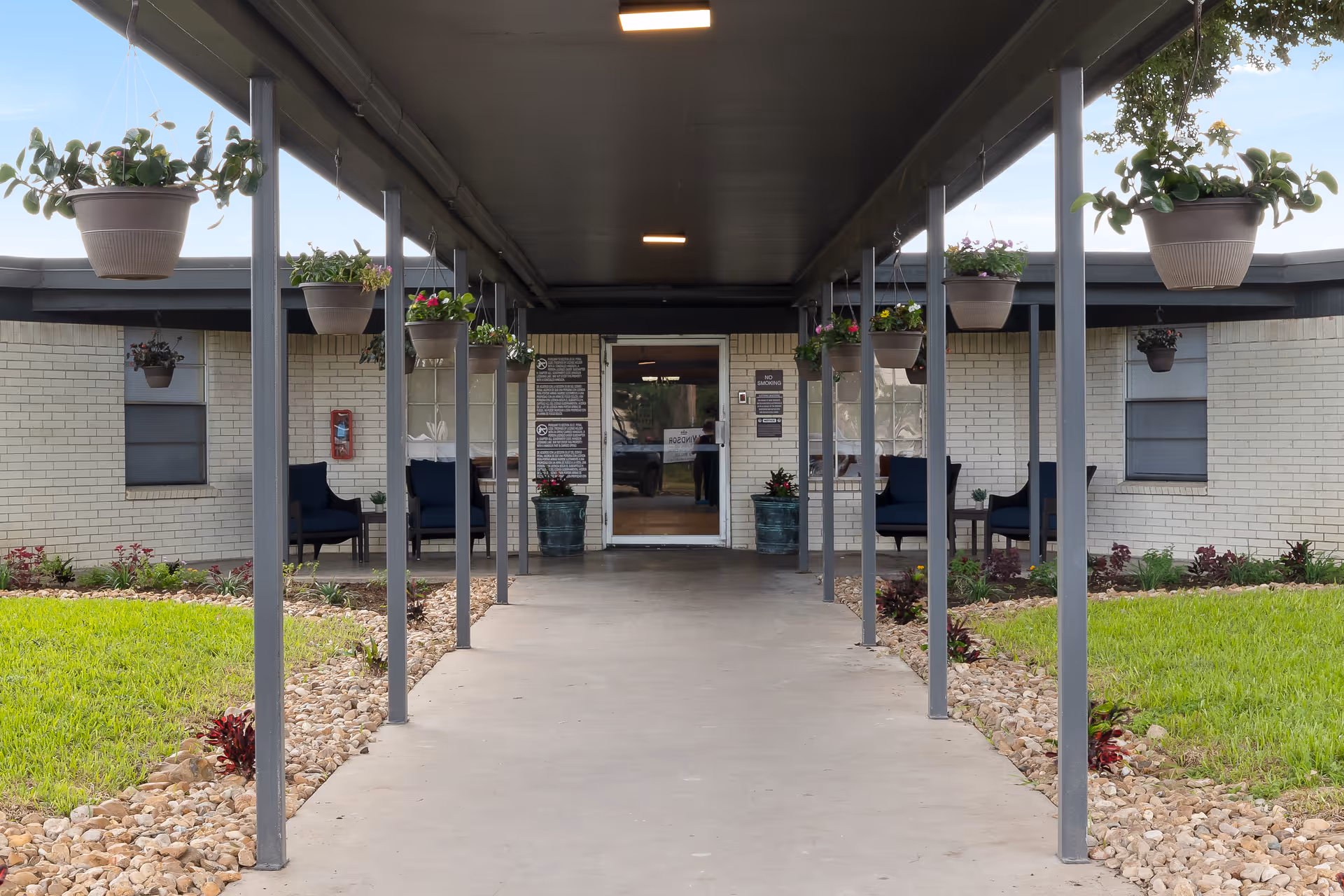Covered walkway leading to the entrance of a building with hanging flower pots on both sides. The building has light-colored brick walls, windows, and seating areas with chairs and tables near the entrance. There are plants and grass along the sides of the walkway.