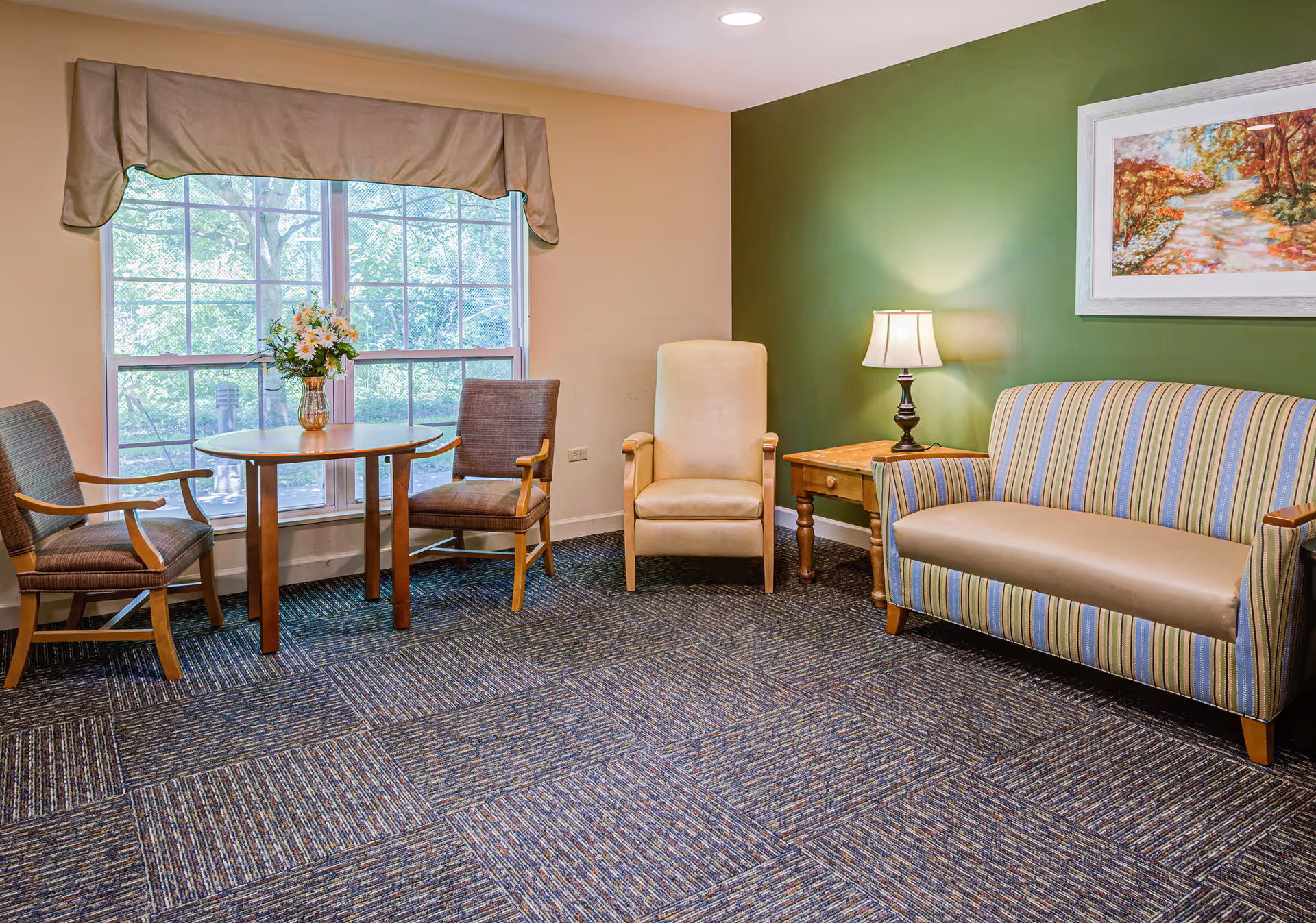 A cozy sitting area with a large window covered by a beige valance. In front of the window, there is a round wooden table with a vase of flowers and two wooden chairs with cushions. To the right, there is a beige armchair next to a wooden side table with a lamp. A striped sofa with blue, green, and beige colors is placed against a green accent wall with a framed painting of a colorful autumn landscape above it. The floor is carpeted with a patterned design.