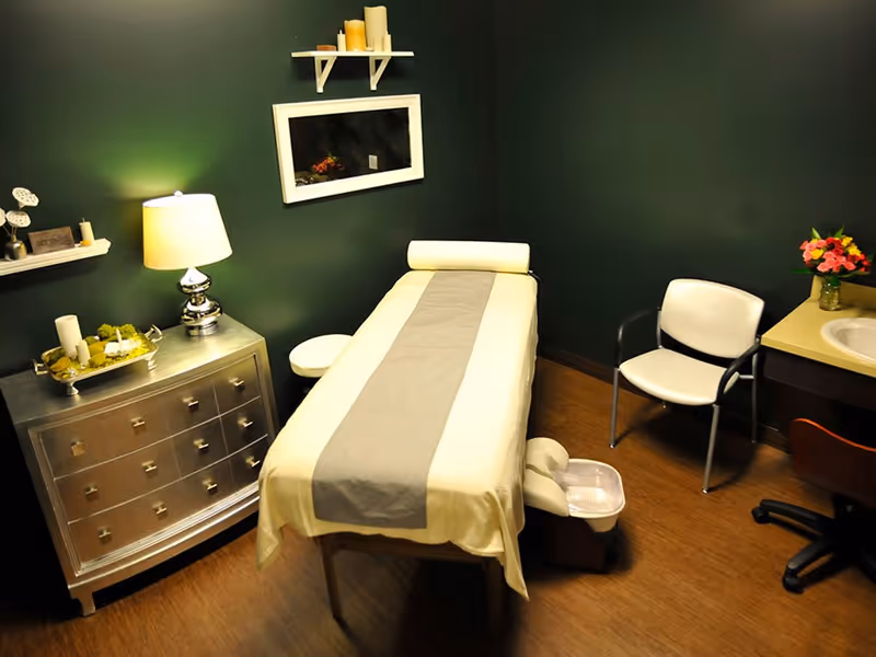 A small treatment room with a massage or therapy table covered with a white sheet and gray runner. The room has dark green walls, a silver chest of drawers with a lamp and decorative items, a white chair, a small sink with a countertop holding a vase of flowers, and a small foot bath near the table.