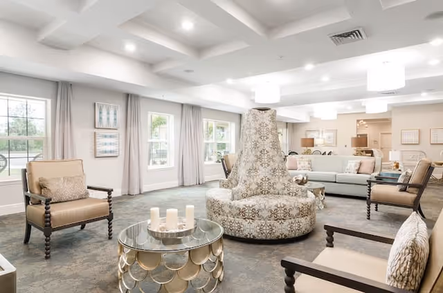 A bright and spacious living room area in a senior living facility featuring multiple armchairs with cushions, a large patterned circular sofa in the center, a round glass coffee table with decorative candles, large windows with curtains allowing natural light, and soft neutral-colored walls and carpet.