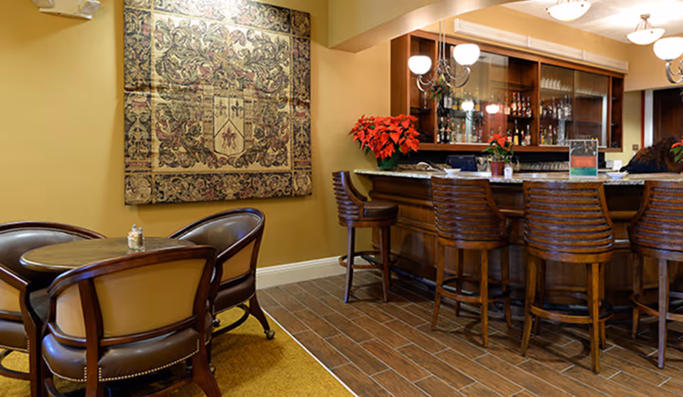 Interior view of a retirement community lounge area featuring a wooden bar with several high chairs, a small round table with four cushioned chairs, a decorative tapestry on the wall, and poinsettia plants placed on the bar counter.