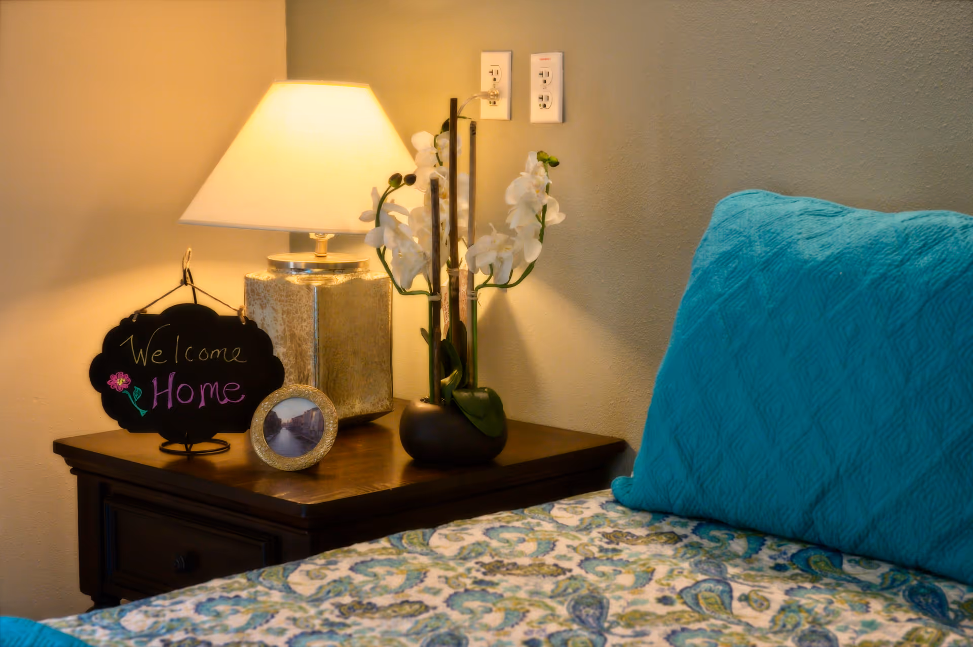 Nightstand with a lamp, potted orchid, picture frame and a 'Welcome Home' sign beside a bed with a teal pillow and patterned bedding.