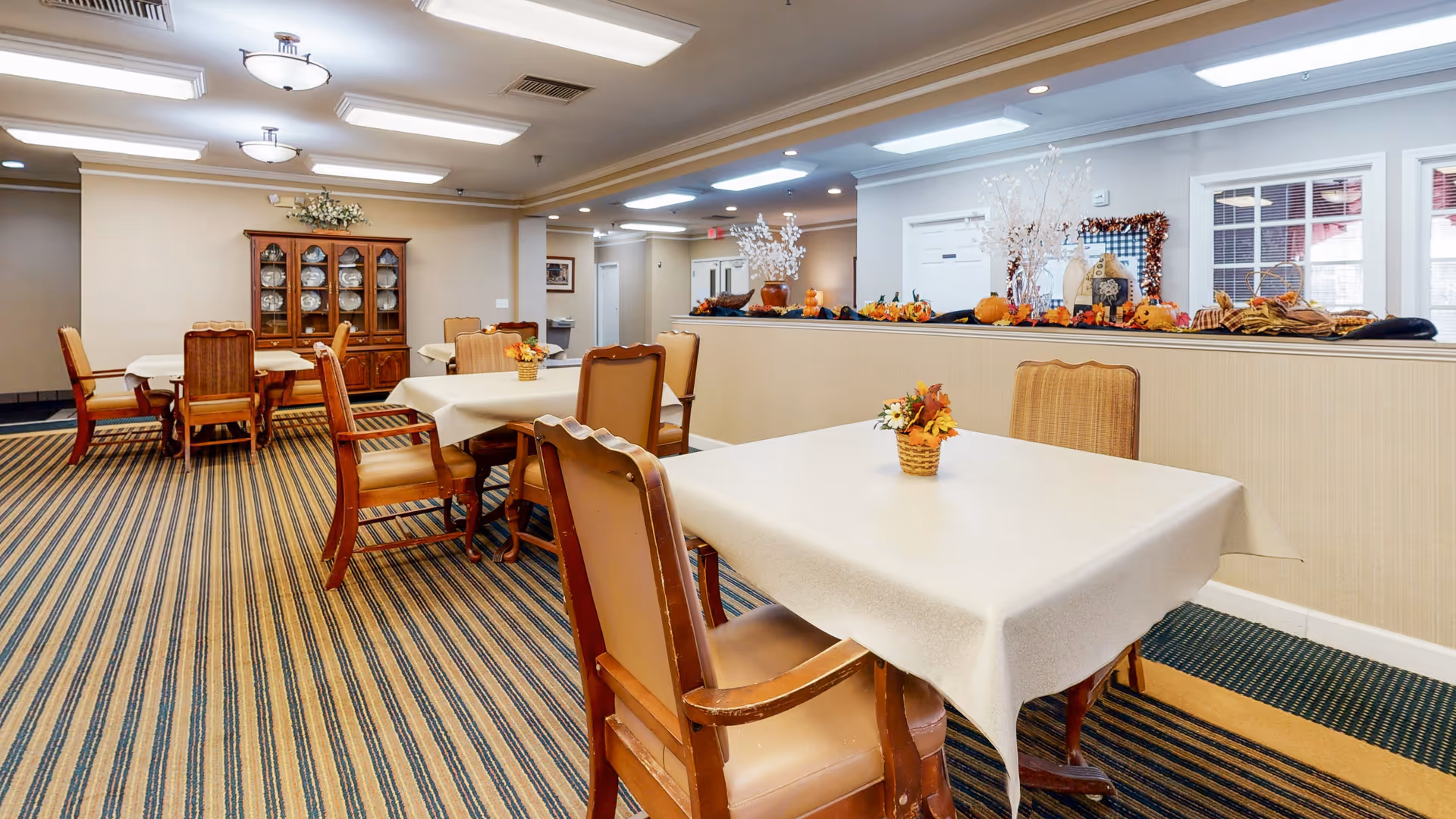 Bright communal dining room with tables draped in white tablecloths, wooden chairs, a china cabinet, and seasonal decorations.