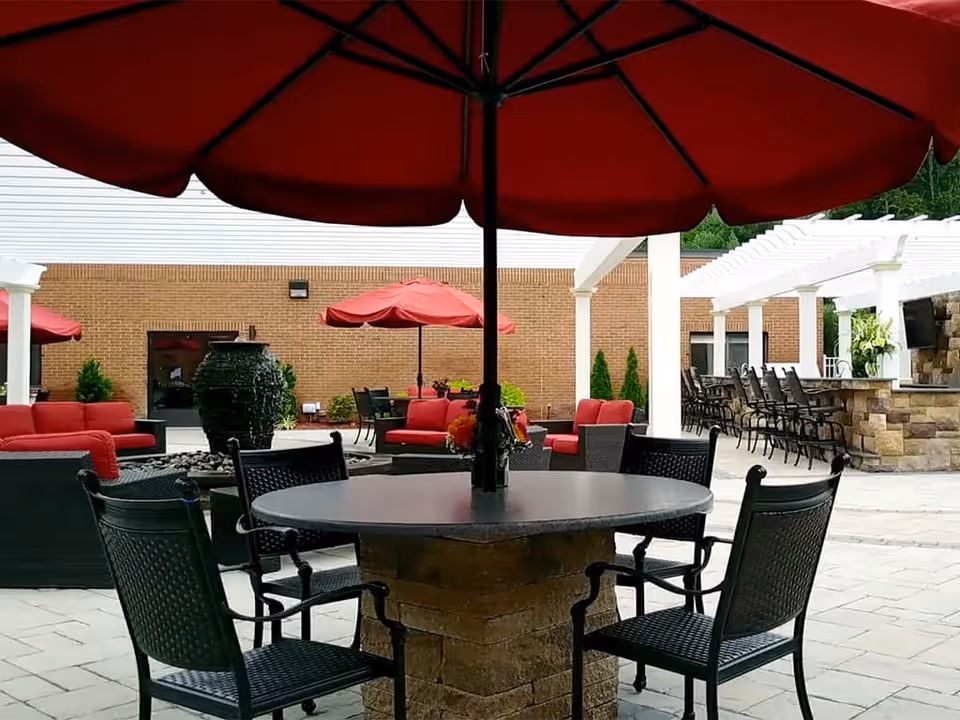 Outdoor patio area with a round table and four black chairs under a large red umbrella. In the background, there are red cushioned seating areas, another red umbrella, a stone water fountain, and a bar area with high chairs under a white pergola. The setting is paved with light-colored tiles and surrounded by a brick building wall.