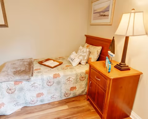 A cozy bedroom with a single bed covered in a seashell-patterned quilt, a gray blanket folded at the foot, and decorative pillows including one with the word 'Blessed'. Next to the bed is a wooden nightstand with a lamp, a small sailboat decoration, and a framed picture hanging on the wall above the bed.