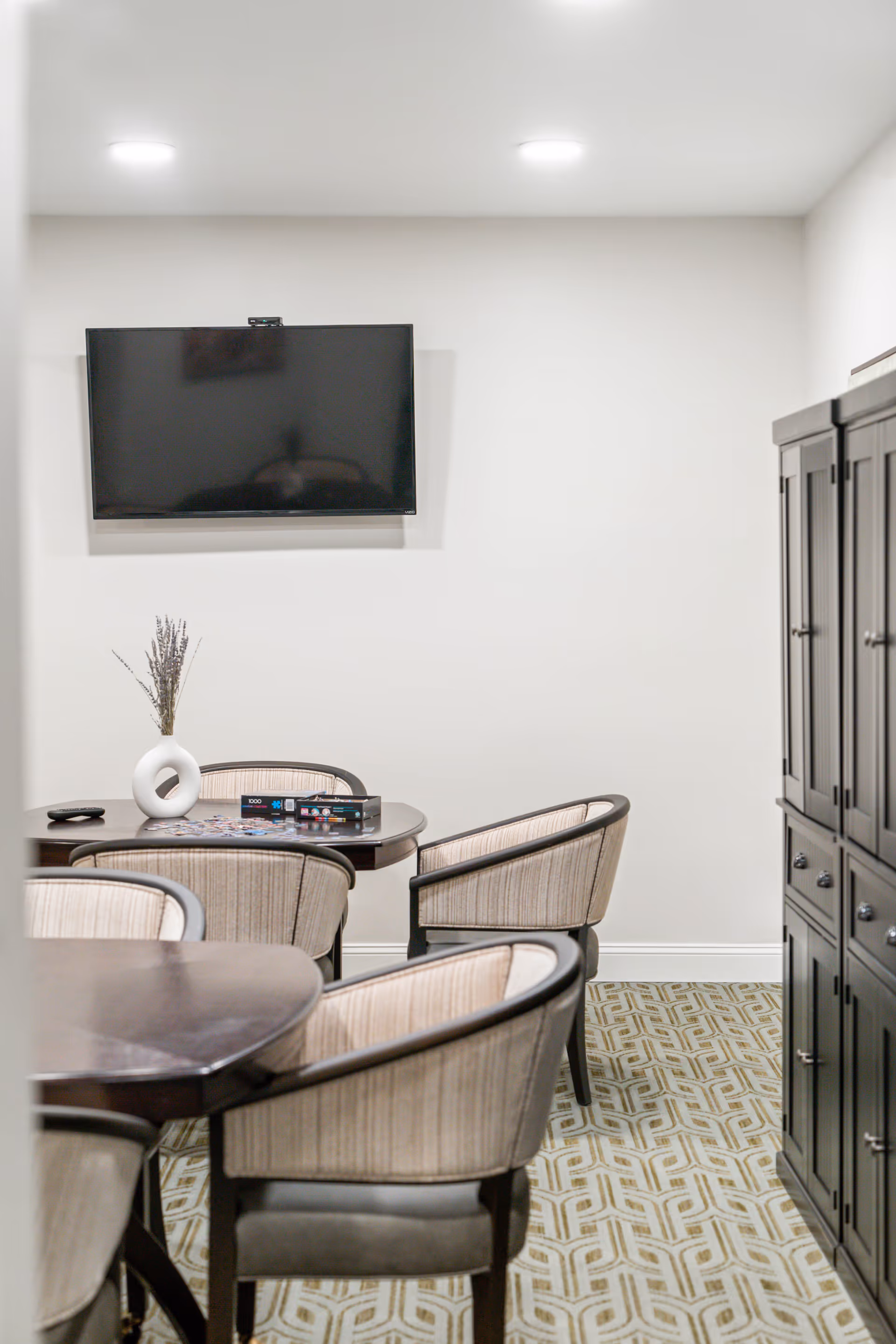Small communal room with round tables and upholstered chairs, a wall-mounted TV, and a storage cabinet.