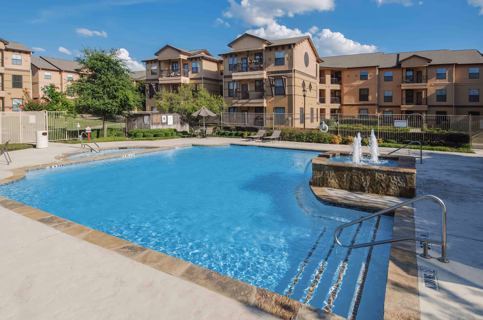 Outdoor swimming pool with clear blue water and a small fountain feature on one side, surrounded by a concrete deck with lounge chairs and umbrellas. In the background, there are multi-story residential buildings with balconies and greenery under a blue sky with some clouds.