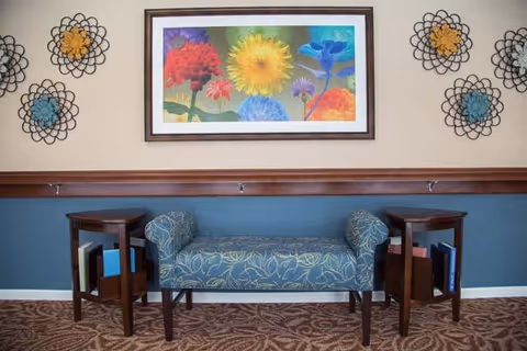 A seating area with a blue patterned bench flanked by two small wooden side tables holding books. Above the bench is a colorful framed painting of various flowers. The wall is decorated with six metal flower-shaped wall hangings in blue, yellow, and black. The lower half of the wall is painted blue, and the upper half is beige. The floor is carpeted with a brown patterned design.
