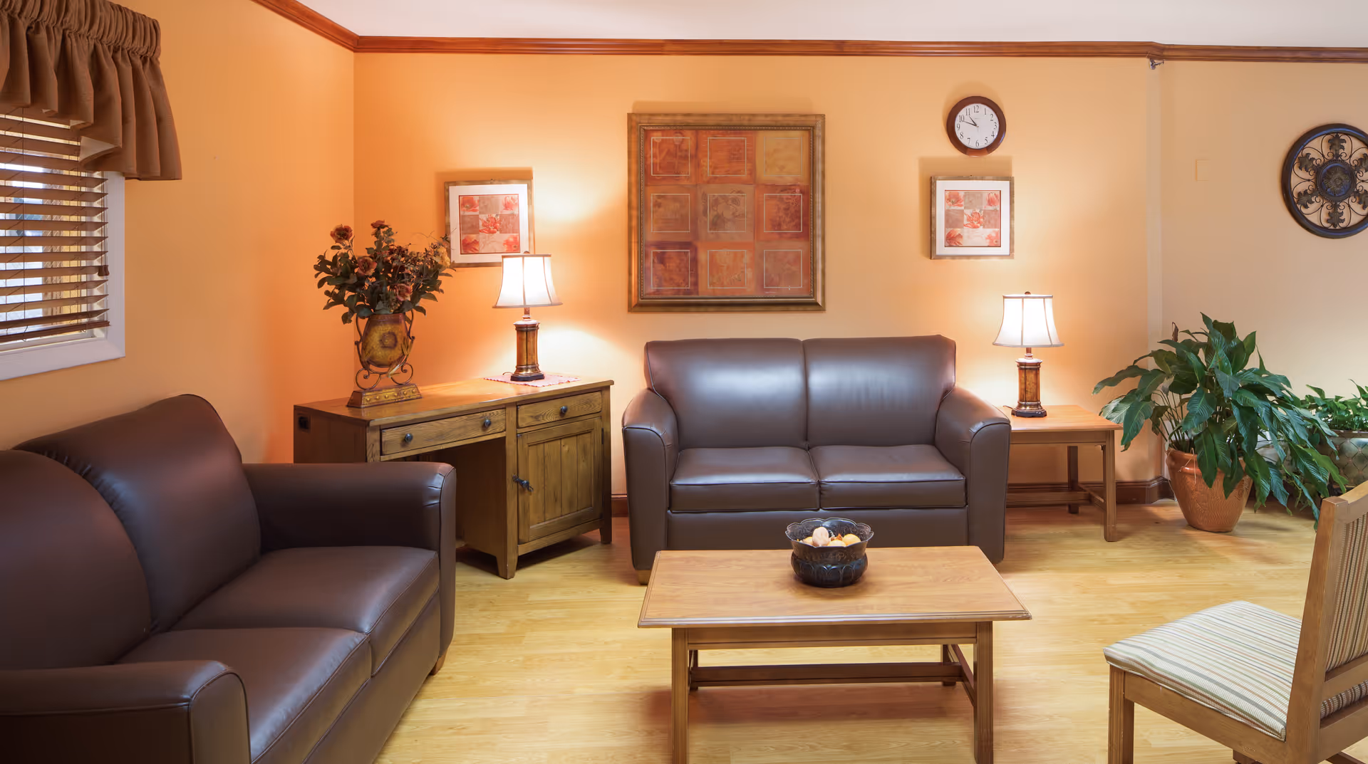 A cozy living room with two brown leather sofas, a wooden coffee table with a decorative bowl, two wooden side tables each with a lamp, a wooden cabinet with a vase of flowers, framed artwork on the peach-colored walls, a clock, and a large potted plant in the corner. The room has wooden flooring and a window with brown blinds and a valance.