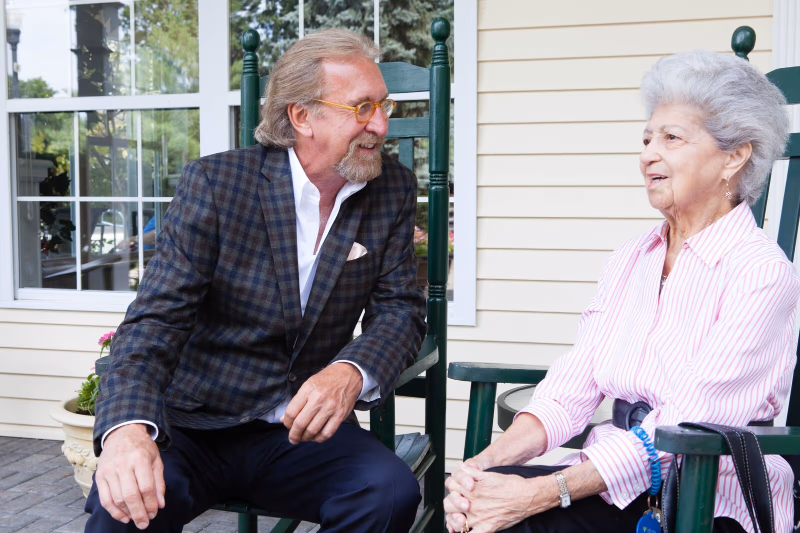 An elderly woman and a middle-aged man sitting on green rocking chairs outside a building with beige siding and a window, engaged in conversation.
