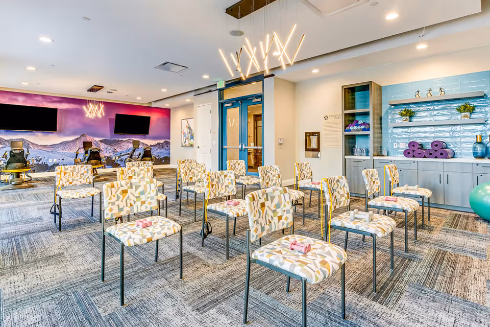 A bright and spacious exercise room with patterned chairs arranged in rows, each chair holding a pair of small dumbbells. The room features a modern light fixture on the ceiling, a wall with a scenic mountain mural and two large flat-screen TVs, and a cabinet area with shelves holding purple yoga mats and plants. Exercise equipment and a large green exercise ball are also visible.