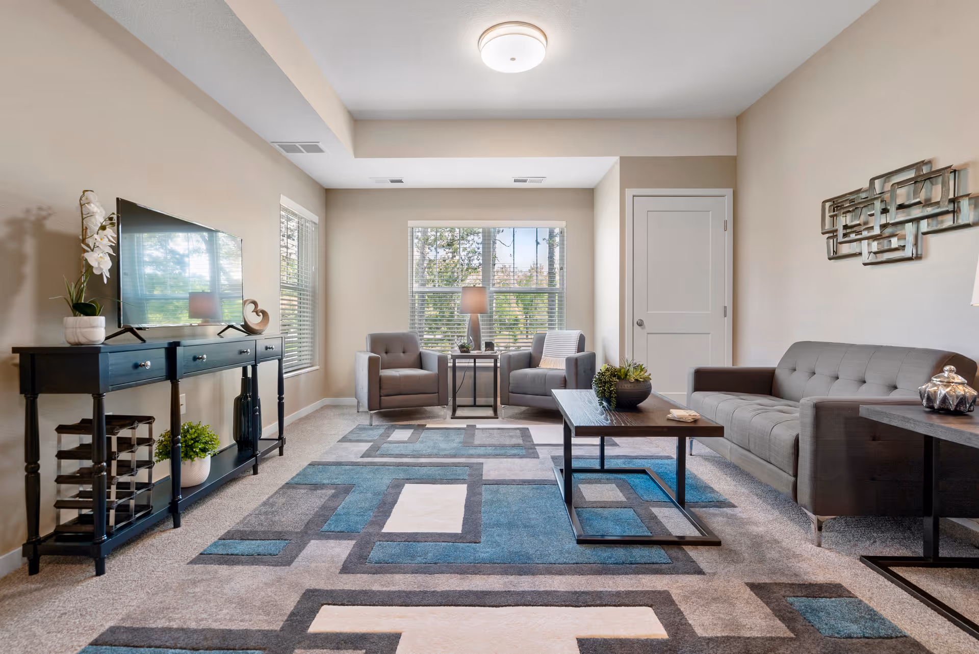 A bright and modern living room with a gray sofa, two gray armchairs, a wooden coffee table, and a patterned area rug in shades of blue, gray, and white. There is a black console table with a flat-screen TV on top, a potted plant, and decorative items. Large windows let in natural light, and a decorative metal wall art piece hangs above the sofa.