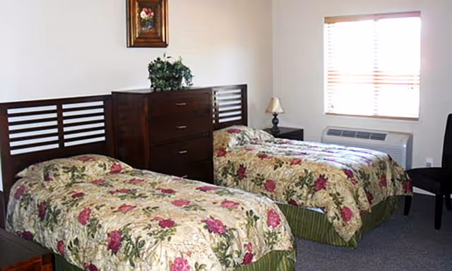 A bedroom with two twin beds featuring floral bedspreads with pink and green patterns. Between the beds is a dark wooden chest of drawers with a green plant on top. A small lamp is on a nightstand next to one bed. A window with blinds allows natural light into the room, and a framed picture hangs on the wall above the chest of drawers.