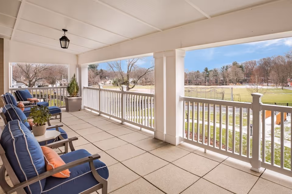Covered outdoor balcony with cushioned blue chairs and small tables, overlooking a grassy field and trees under a blue sky.
