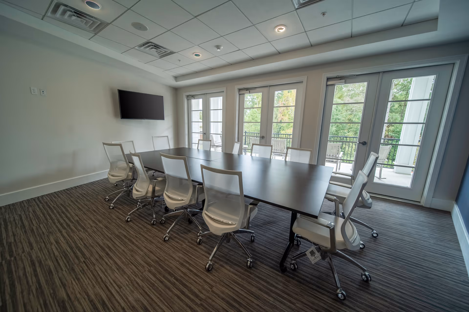 A modern conference room with a long rectangular table surrounded by white office chairs on wheels. The room has large windows and glass doors that open to an outdoor balcony with seating. A flat-screen TV is mounted on the wall opposite the windows. The floor is carpeted with a striped pattern, and the ceiling has recessed lighting and ventilation panels.
