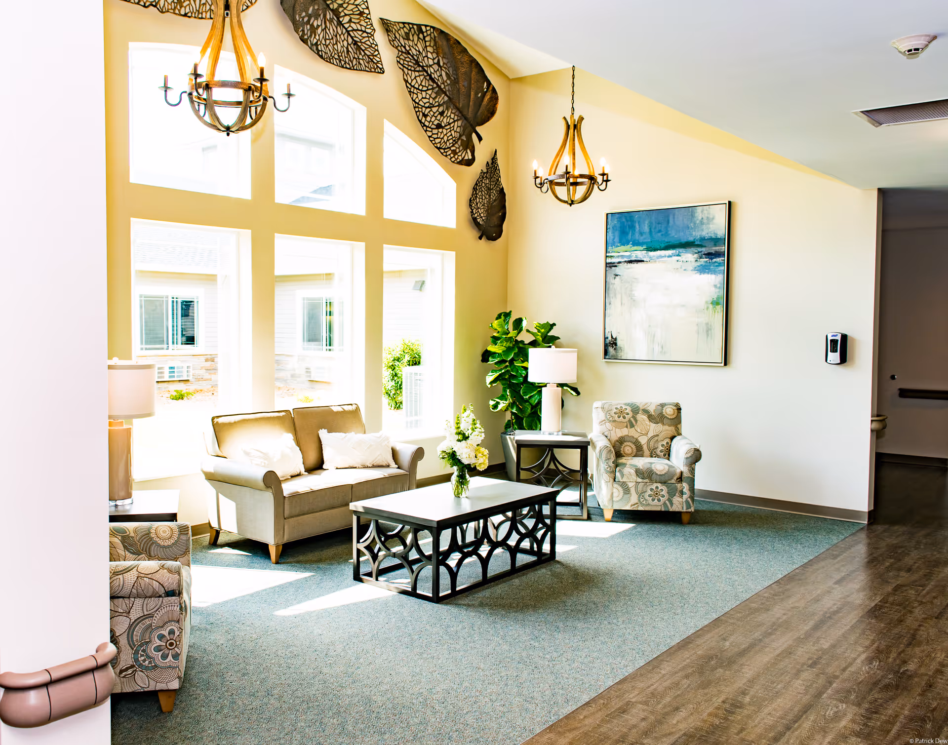 A bright and cozy living room area with large windows letting in natural light. The room features a beige sofa with two white pillows, a patterned armchair, a dark wooden coffee table with a vase of flowers, and a side table with a lamp. The walls are decorated with large metal leaf sculptures and a framed abstract painting. Two chandeliers hang from the ceiling, and there is a green plant in the corner.