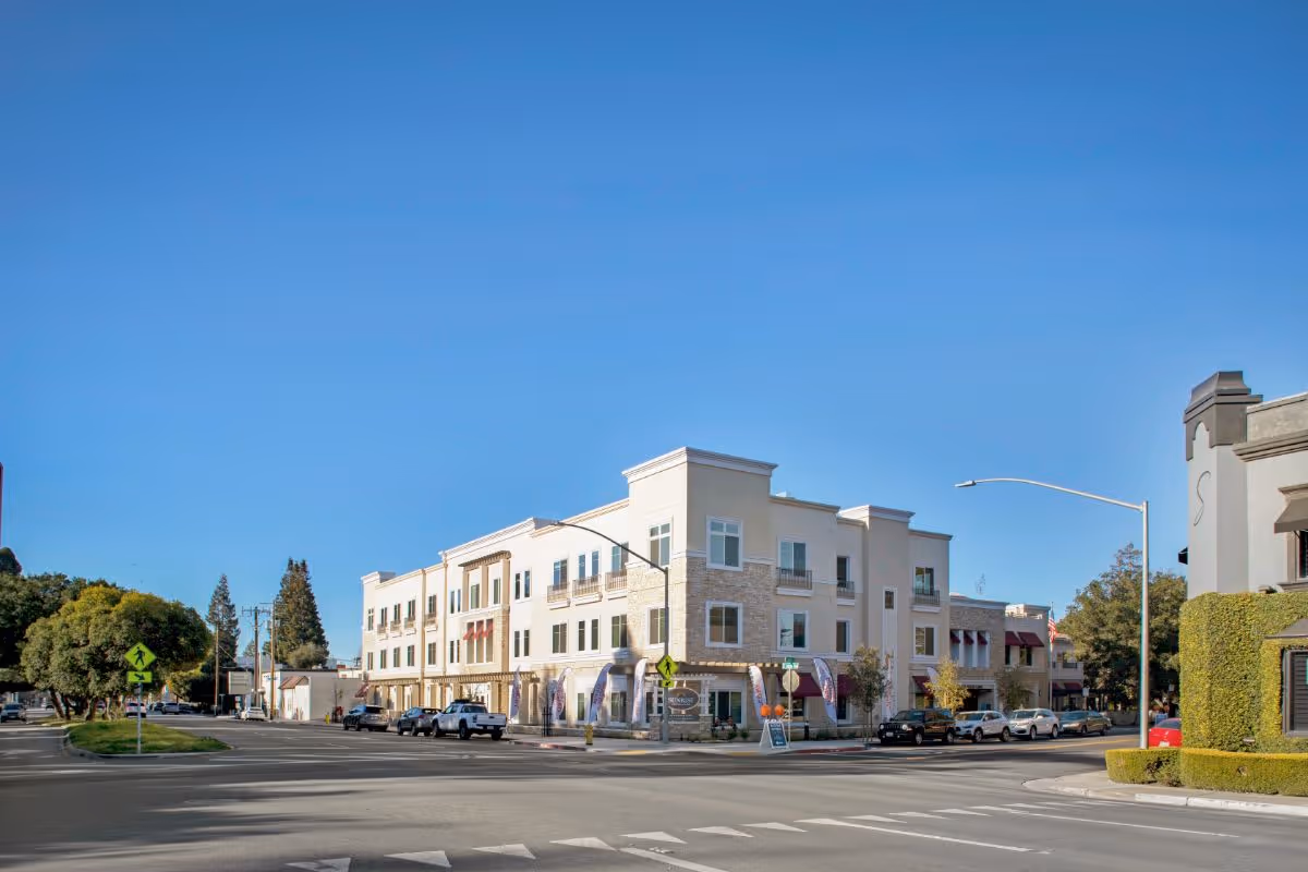 Exterior view of a three-story building on a street corner under a clear blue sky. The building has a beige and stone facade with multiple windows and balconies. Several cars are parked along the street, and there are trees and street signs visible nearby.