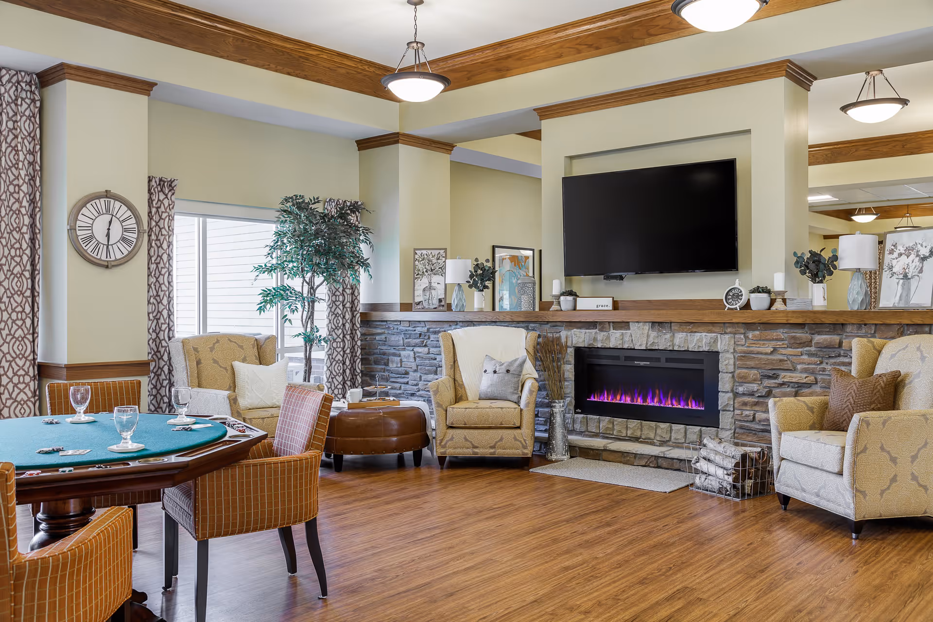 A cozy living room area with a stone fireplace featuring a modern electric fire with purple flames. Above the fireplace is a mounted flat-screen TV. The room has wooden flooring and crown molding with recessed lighting. There are comfortable armchairs with cushions, a round table with chairs set for a game, a large potted plant, and decorative items on the mantel and walls.