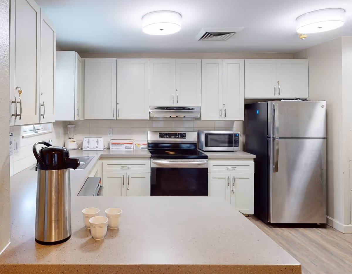 A modern kitchen with white cabinets, a stainless steel refrigerator, stove, and microwave. There is a countertop with a large silver coffee dispenser and three white disposable cups. The kitchen has bright overhead lighting and a light-colored floor.