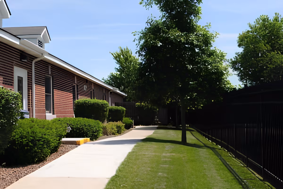 Outdoor walkway alongside a brick building with neatly trimmed bushes and trees on a sunny day. A black metal fence runs parallel to the walkway on the right side, with green grass between the path and the fence.