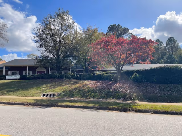 Exterior view of Lawndale Manor Assisted Living facility with a lawn, trees including one with red leaves, and a clear blue sky with some clouds.