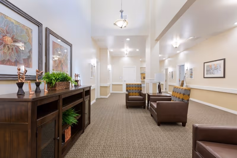 A well-lit interior hallway or common area in a senior living facility with beige walls and carpeted floor. There are two brown armchairs with patterned cushions, a wooden sideboard with decorative items including small sculptures and a potted plant, and framed artwork on the walls. Ceiling lights provide warm illumination.