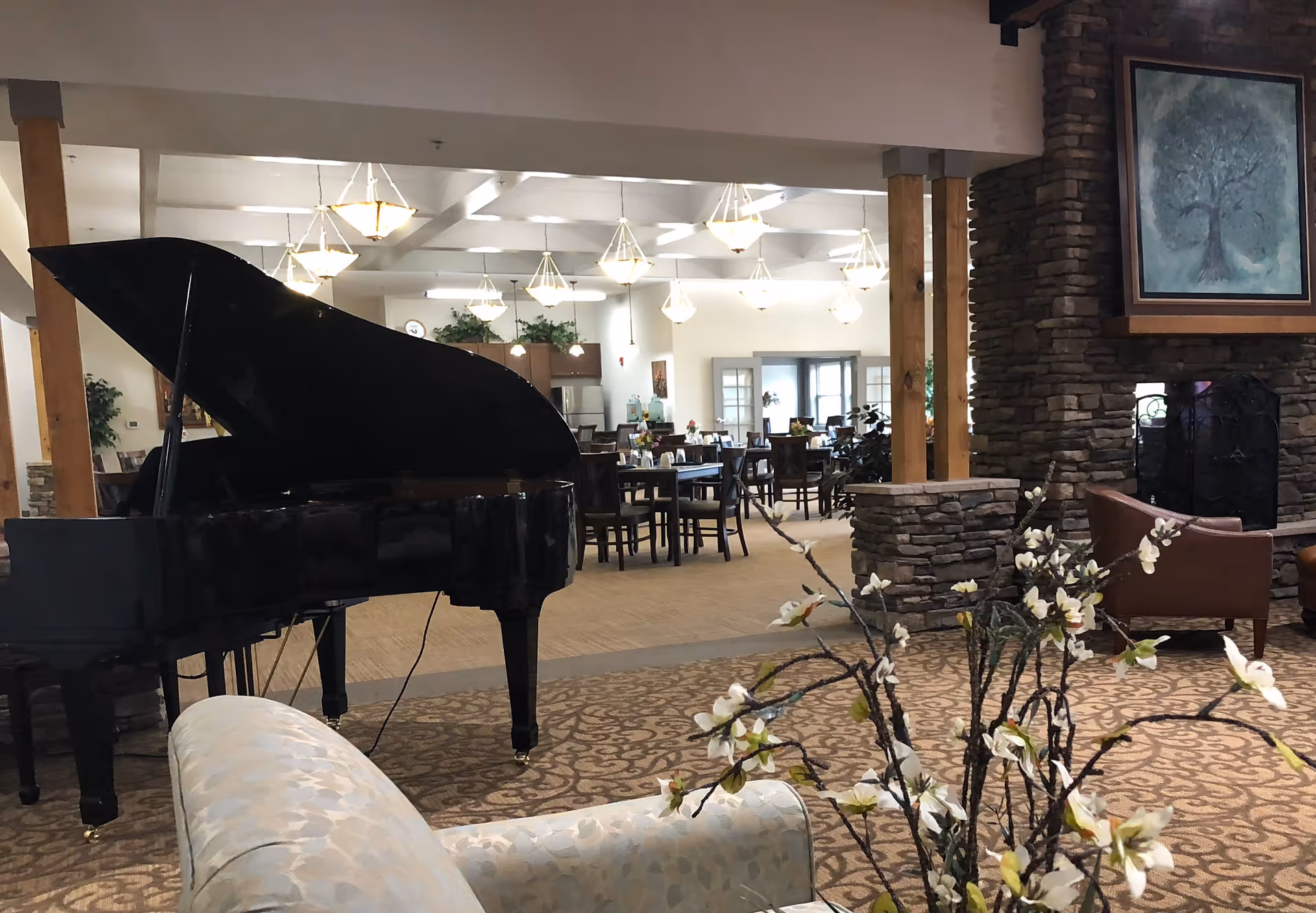 Interior view of a senior living facility showing a black grand piano, a patterned armchair, a stone fireplace with a painting above it, and a dining area with tables and chairs in the background. The space is well-lit with hanging light fixtures and decorated with plants and flowers.