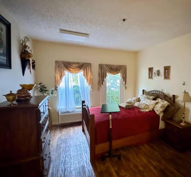 A cozy bedroom with a wooden bed covered in a red blanket and multiple pillows. There are two windows with sheer white curtains and decorative valances letting in natural light. A wooden dresser with decorative items is on the left side, and a nightstand with a lamp is on the right side of the bed. The floor is wood, and the walls are painted a light color.