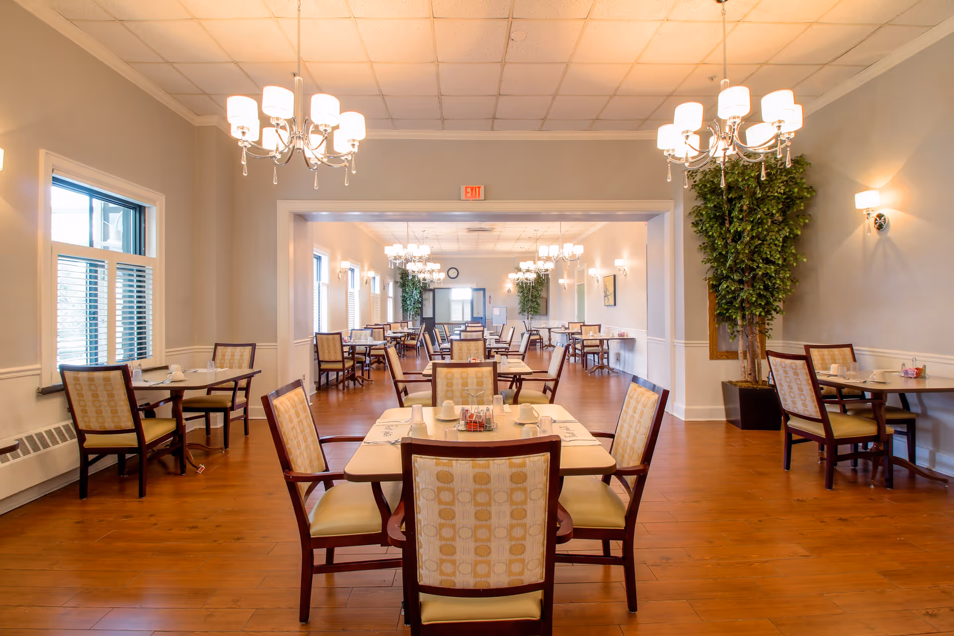 A spacious and well-lit dining room in a senior living facility with multiple tables and chairs arranged neatly. The room features wooden flooring, large windows with blinds, elegant chandeliers, and potted plants along the walls. Tables are set with cups, plates, and condiments.