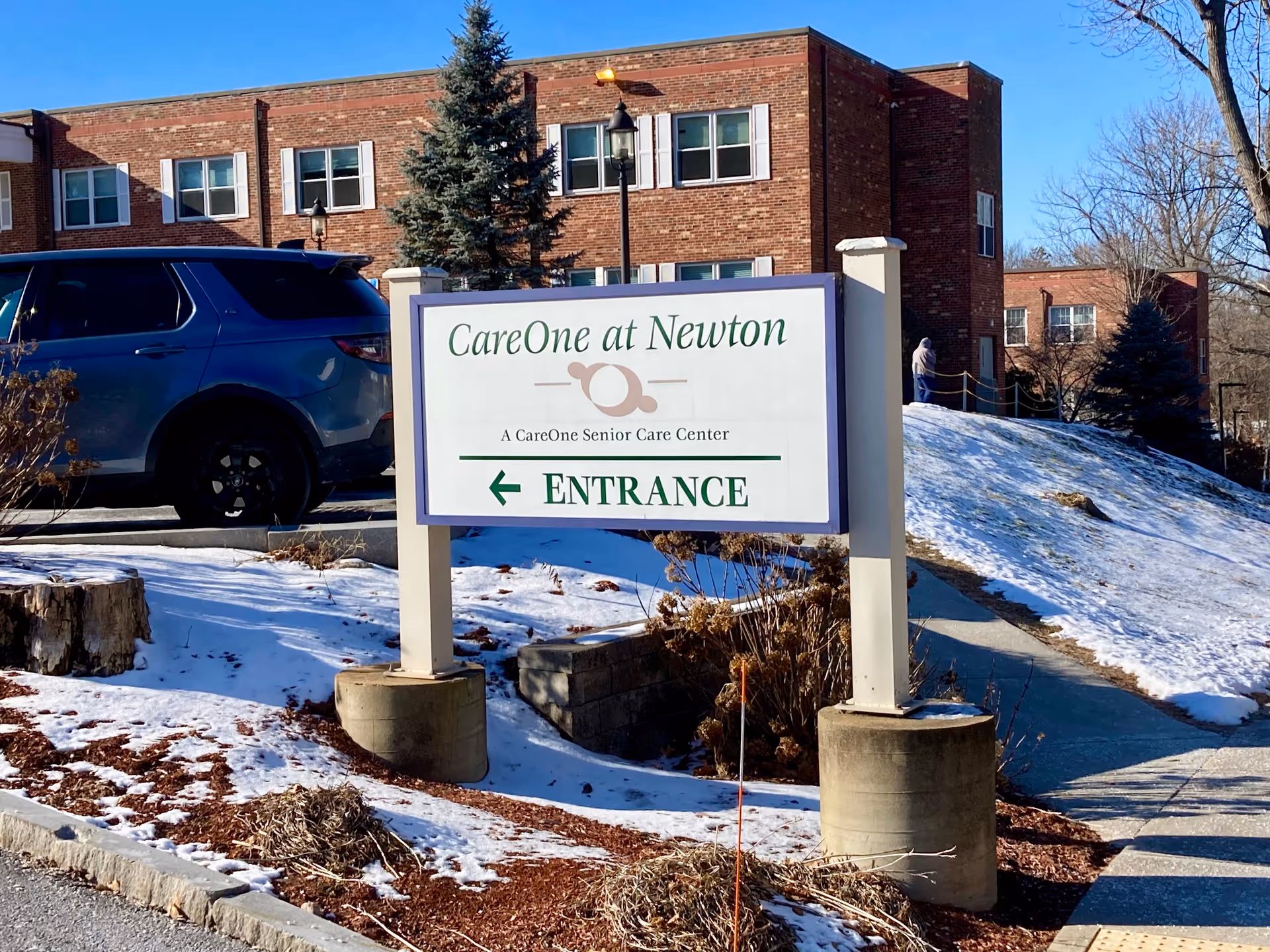 Outdoor view of CareOne at Newton senior care center sign with a brick building and a parked blue SUV in the background. Snow covers the ground and a person is walking on a path near the building.
