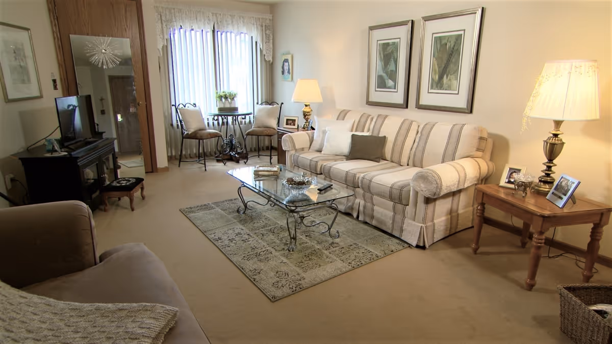 A cozy living room with a striped sofa, glass coffee table on a patterned rug, a side table with a lamp and framed photos, two chairs and a small round table near a window with vertical blinds, a TV on a stand, and a large mirror on the wall.