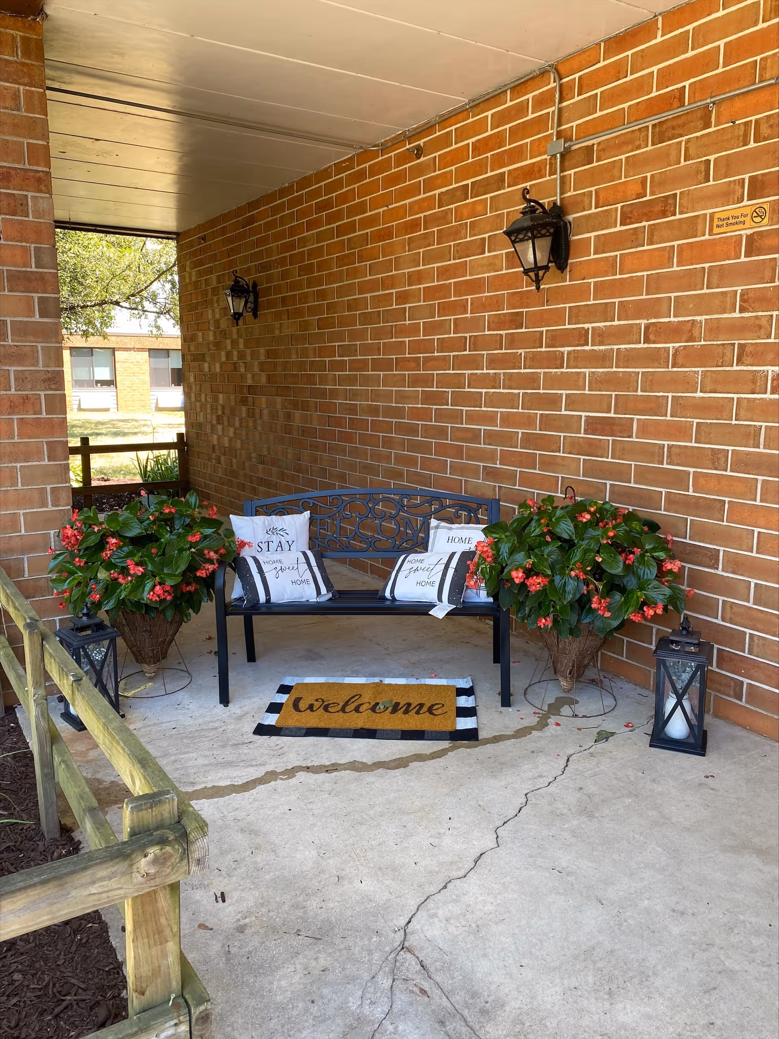 A covered outdoor seating area with a black metal bench adorned with three decorative pillows that have welcoming messages. Two large potted plants with green leaves and red flowers flank the bench. A welcome mat is placed on the concrete floor in front of the bench. Two black lanterns with candles inside are positioned near the plants. The background features a brick wall with two mounted lantern-style lights and a small sign that reads 'Thank You For Not Smoking'.